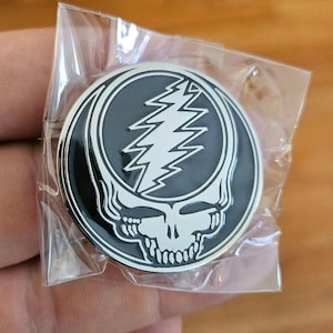 May include: A round, silver-rimmed pin featuring a black skull with a white lightning bolt design. The pin is in a clear plastic package. The skull has a stylized design with a wide grin and a lightning bolt in the center.
