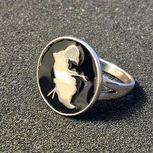 May include: Silver ring with a black oval face featuring a white silhouette of a person holding a bow and arrow. The ring has a split band design and is set against a dark, textured background.