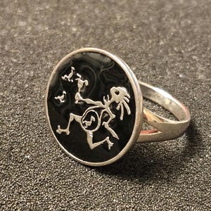 May include: Silver ring with a black oval face featuring a stylized figure with a circular design on its torso. The ring has a split band and a detailed design.