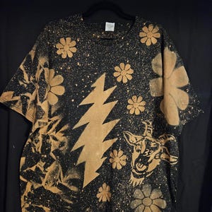 May include: Black t-shirt with a bleach-dyed design featuring a lightning bolt, flowers, and a goat graphic. The shirt has a splatter effect and is hanging on a black hanger.