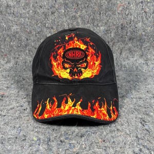 Puede incluir: Gorra de b&eacute;isbol negra con un gr&aacute;fico de calavera en llamas y el texto "NHRA". El dise&ntilde;o de la gorra presenta llamas rojas y amarillas alrededor de la calavera y en la visera. La gorra est&aacute; hecha de tela duradera.