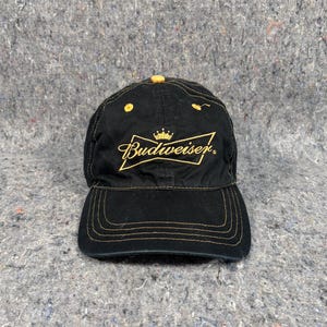 May include: Black baseball cap featuring gold stitching and the Budweiser logo in gold script. The cap has a curved peak and yellow accents. A classic, casual accessory.