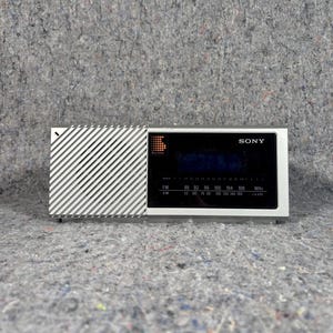 May include: A rectangular Sony radio featuring a silver-toned speaker grille with diagonal slats on the left side and a black control panel on the right. The panel displays FM and AM frequency scales, a digital-style logo, and the Sony brand name.