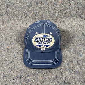 May include: A navy blue baseball cap with the text "MAPLE LEAFS TORONTO" in white and blue. The cap has a curved brim and white stitching details. The Maple Leafs logo is centered on the front.