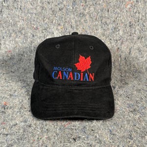 以下が含まれることがあります： 黒の野球帽で、青い文字の「MOLSON」と赤い文字の「CANADIAN」の上に、赤いカエデの葉が刺繍されています。この帽子は構造化されたクラウン、カーブしたつば、そして通気のための上部パネルのアイレットを備えており、クラシックな外観を作り出しています。