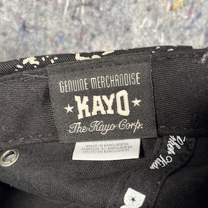 Może przedstawiać: Wnętrze czarnej czapki z prostokątną tkaną metką z napisem GENUINE MERCHANDISE KAYO The Kayo Corp. Mniejsza biała metka poniżej zawiera napis MADE IN BANGLADESH. Wewnętrzna krawędź posiada biały napis DGK oraz inne częściowe litery.