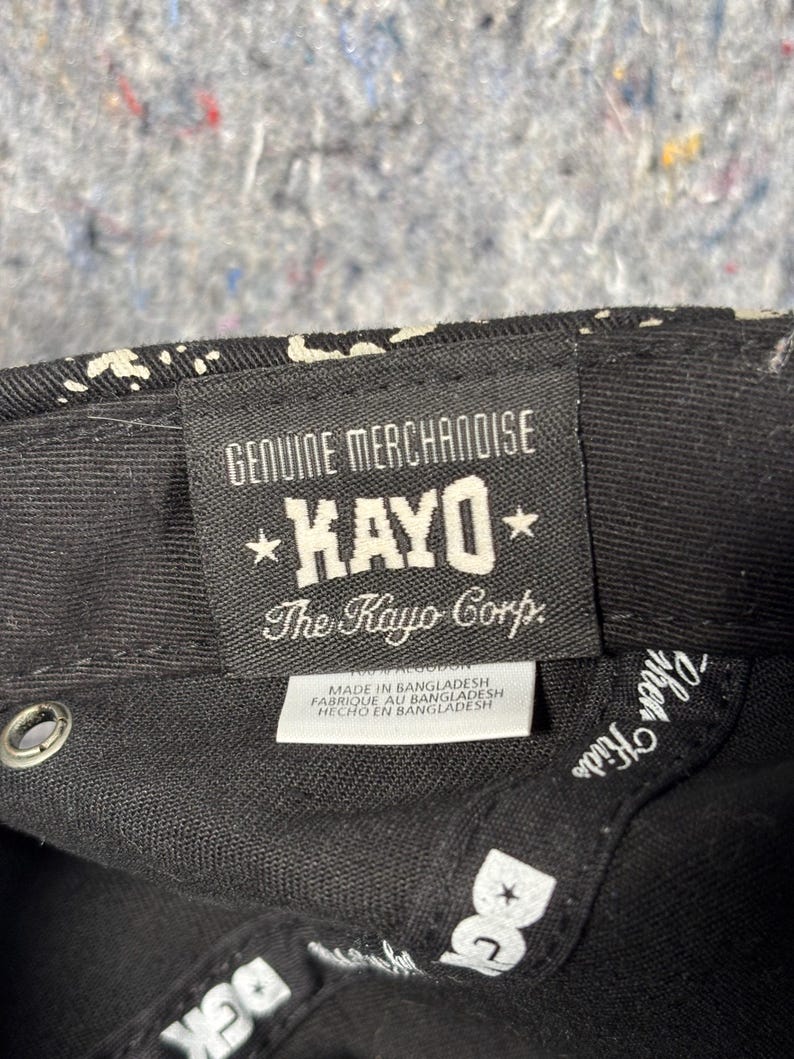 Może przedstawiać: Wnętrze czarnej czapki z prostokątną tkaną metką z napisem GENUINE MERCHANDISE KAYO The Kayo Corp. Mniejsza biała metka poniżej zawiera napis MADE IN BANGLADESH. Wewnętrzna krawędź posiada biały napis DGK oraz inne częściowe litery.