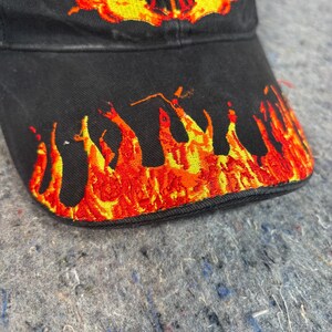Puede incluir: Gorra de b&eacute;isbol negra con llamas bordadas y un dise&ntilde;o de calavera en tonos rojos, naranjas y amarillos. Las llamas est&aacute;n en el borde y la calavera est&aacute; centrada en la gorra.