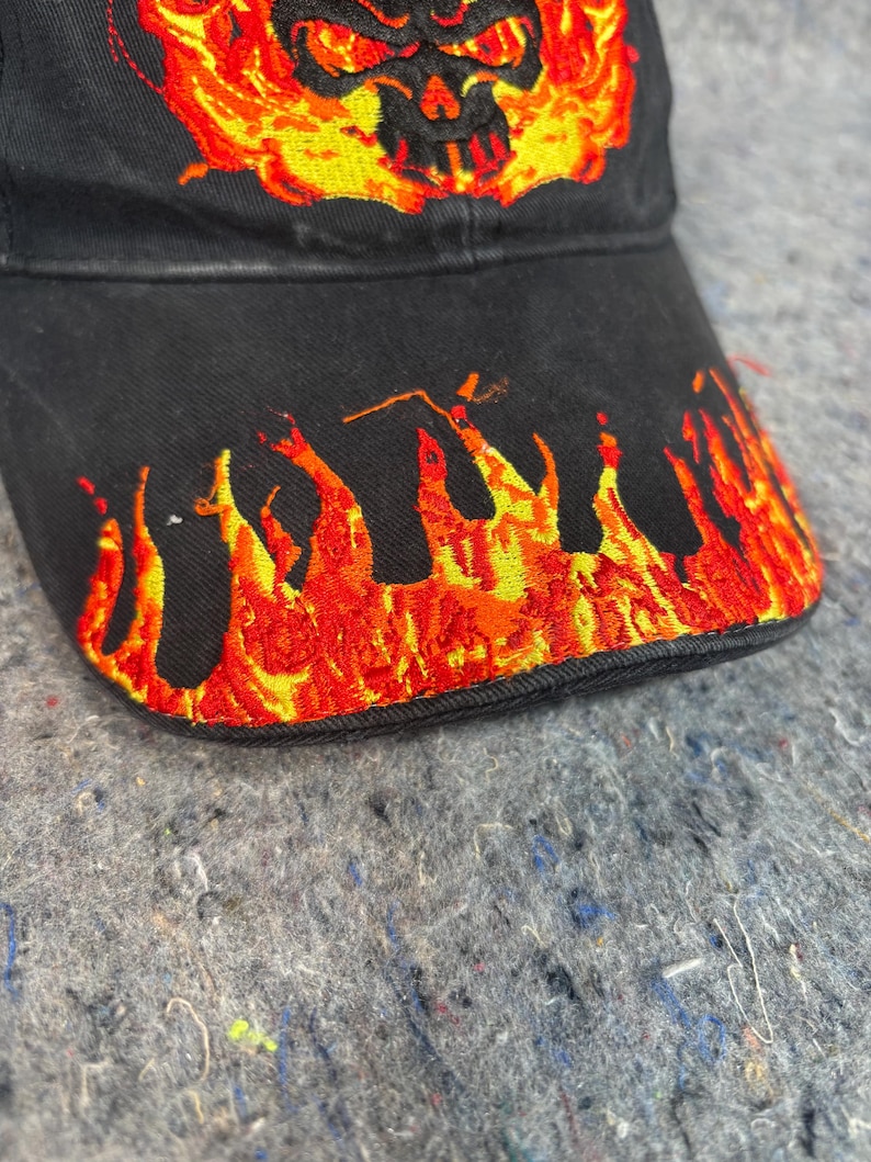 Puede incluir: Gorra de b&eacute;isbol negra con llamas bordadas y un dise&ntilde;o de calavera en tonos rojos, naranjas y amarillos. Las llamas est&aacute;n en el borde y la calavera est&aacute; centrada en la gorra.