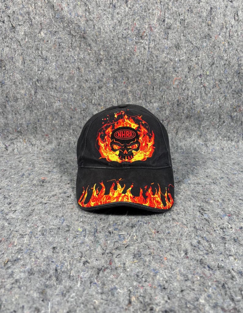 Puede incluir: Gorra de b&eacute;isbol negra con un gr&aacute;fico de calavera en llamas y el texto "NHRA". El dise&ntilde;o de la gorra presenta llamas rojas y amarillas alrededor de la calavera y en la visera. La gorra est&aacute; hecha de tela duradera.