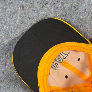 May include: A yellow and black baseball cap with the text "TAG" visible on the inside. The cap has a black peak and a yellow crown. The interior is tan with black stitching and ventilation holes.