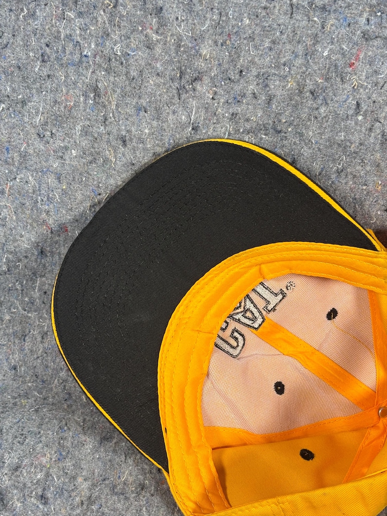 May include: A yellow and black baseball cap with the text "TAG" visible on the inside. The cap has a black peak and a yellow crown. The interior is tan with black stitching and ventilation holes.