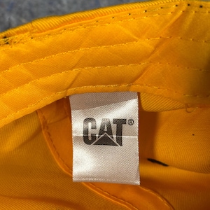 May include: Close-up of the inside of a yellow baseball cap. A white tag features the black "CAT" logo. The cap's interior is bright yellow, with visible stitching and a black button.