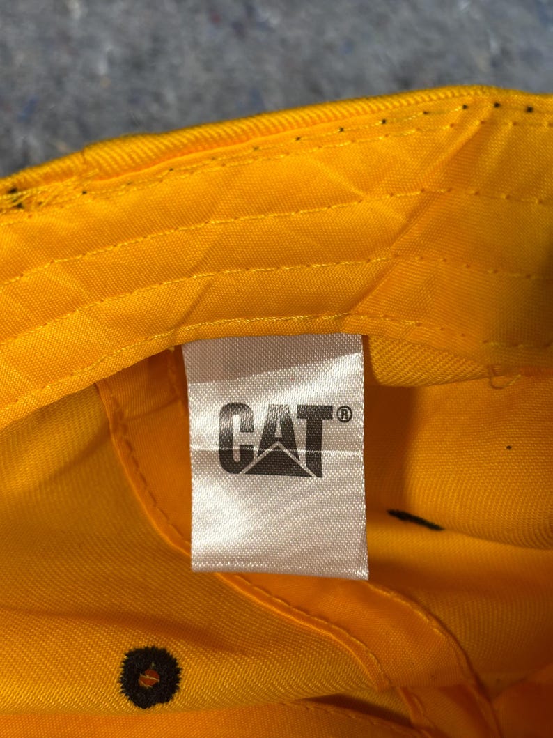 May include: Close-up of the inside of a yellow baseball cap. A white tag features the black "CAT" logo. The cap's interior is bright yellow, with visible stitching and a black button.