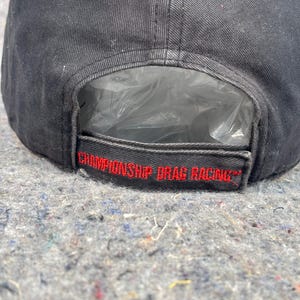 Puede incluir: Gorra de b&eacute;isbol negra con el texto "CHAMPIONSHIP DRAG RACING" bordado en rojo. La gorra tiene una visera curva y una correa de ajuste de pl&aacute;stico. Hecha de tela duradera.