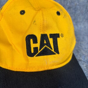 May include: Yellow and black baseball cap featuring the word "CAT" in black block letters and a black logo. The cap has a curved peak and a black button on top.