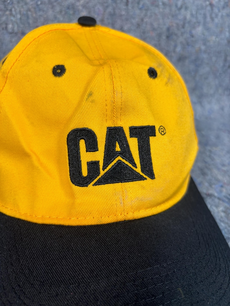 May include: Yellow and black baseball cap featuring the word "CAT" in black block letters and a black logo. The cap has a curved peak and a black button on top.