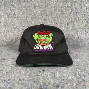 May include: Black baseball cap featuring the Coca-Cola "Monsters of the Gridiron" logo in green, orange, and purple. The cap has a curved bill and a green underside.