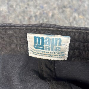 Puede incluir: Primer plano de una gorra de b&eacute;isbol negra con una etiqueta blanca. La etiqueta tiene el texto "main gate" en letras azules. La etiqueta tambi&eacute;n tiene el texto "vintage has an entrance" y "100% algod&oacute;n hecho en China".