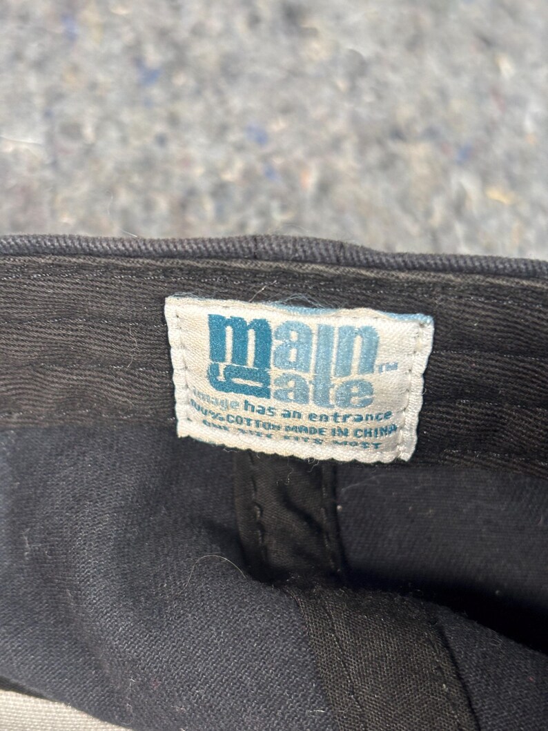 Puede incluir: Primer plano de una gorra de b&eacute;isbol negra con una etiqueta blanca. La etiqueta tiene el texto "main gate" en letras azules. La etiqueta tambi&eacute;n tiene el texto "vintage has an entrance" y "100% algod&oacute;n hecho en China".