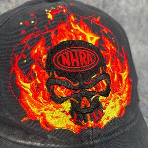 Puede incluir: Gorra de b&eacute;isbol negra con un dise&ntilde;o de calavera bordada rodeada de llamas. La calavera tiene ojos rojos y est&aacute; enmarcada por llamas naranjas y amarillas. El texto "NHRA" est&aacute; bordado en rojo encima de la calavera.