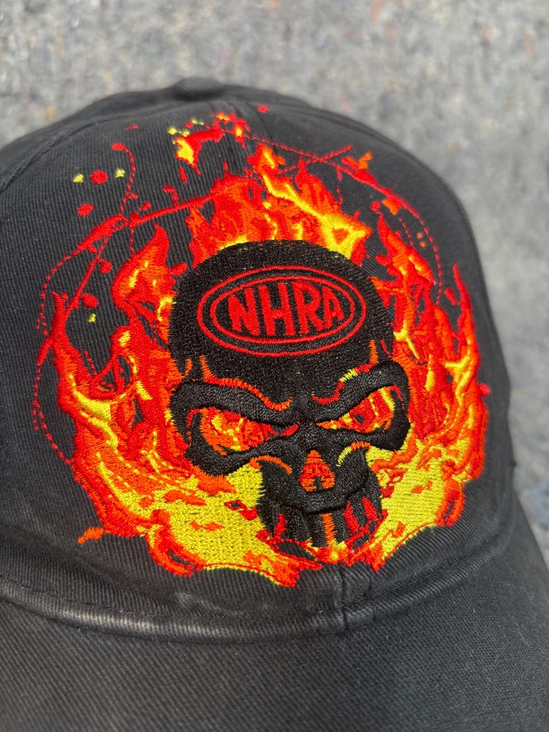 Puede incluir: Gorra de b&eacute;isbol negra con un dise&ntilde;o de calavera bordada rodeada de llamas. La calavera tiene ojos rojos y est&aacute; enmarcada por llamas naranjas y amarillas. El texto "NHRA" est&aacute; bordado en rojo encima de la calavera.