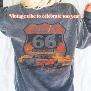May include: Gray sweatshirt featuring a Route 66 graphic with a vintage car and desert landscape. The design includes the text "Vintage vibe to celebrate 100 years" and "66 1926-2026".