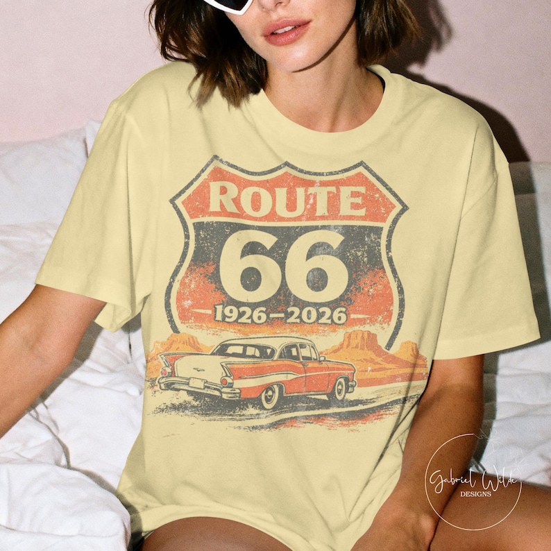 May include: A light yellow t-shirt featuring a Route 66 design. The design includes a red and gray shield with "ROUTE 66" and "66" printed on it, along with the years "1926-2026". Below the text is an illustration of a classic red car.