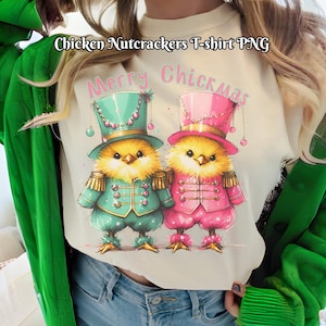 May include: Cream-colored t-shirt featuring two cartoon chicks dressed as nutcrackers. One chick wears a teal outfit and hat, the other a pink outfit and hat. The text "Merry Chickmas" is above the chicks.