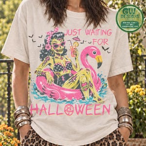 May include: A beige t-shirt featuring a graphic of a zombie pin-up character in a pink flamingo pool float, holding a cocktail. The text reads "JUST WAITING FOR HALLOWEEN" with a pentagram in the "O". Bats and water splashes surround the figure.
