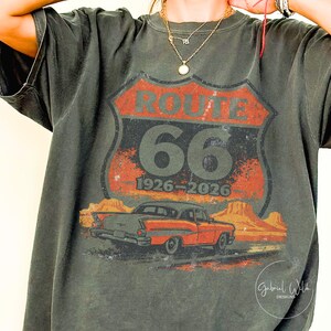 May include: Dark gray t-shirt featuring a Route 66 graphic. The design includes a red and orange shield with "ROUTE 66" and "66" in large numbers, along with the years "1926-2026". A vintage red car is depicted below the text, with a desert landscape in the background.