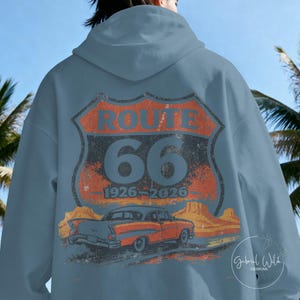 May include: A light blue hoodie featuring a distressed Route 66 design on the back. The design includes the words "ROUTE 66" above the number "66" and the years "1926-2026", along with an illustration of a classic car.