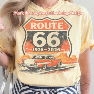 May include: Cream-colored t-shirt featuring a Route 66 design with a vintage car graphic. The design includes the text "ROUTE 66" and the years "1926-2026". The shirt also has the text "Mark the 100 years with vintage design".