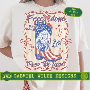 Puede incluir: Camiseta color crema con un diseño patriótico de una bota de vaquero de la Ruta 66. La bota es roja, blanca y azul, con el texto "Freedom Runs This Road". Incluye el texto "GWD GABRIEL WILDE DESIGNS".
