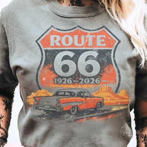 May include: Gray t-shirt featuring a Route 66 design. The design includes a road sign with "ROUTE 66" and "66" in large numbers, along with the years "1926-2026" and an orange classic car.
