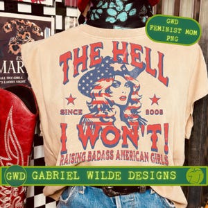 Könnte beinhalten: Beiges T-Shirt mit einem Aufdruck einer Frau mit patriotischem Hut, dem Text "THE HELL I WON'T!" und "RAISING BADASS AMERICAN GIRLS." Das Design enthält Sterne und den Text "SINCE 2005."