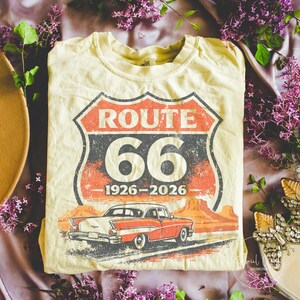 May include: Cream-colored t-shirt featuring a Route 66 design with a vintage car graphic. The design includes the words "ROUTE 66" in red and white, the number "66", and the years "1926-2026". The car is red and white.