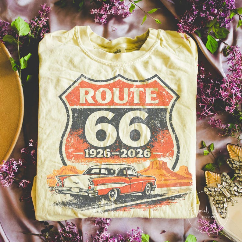 May include: Cream-colored t-shirt featuring a Route 66 design with a vintage car graphic. The design includes the words "ROUTE 66" in red and white, the number "66", and the years "1926-2026". The car is red and white.