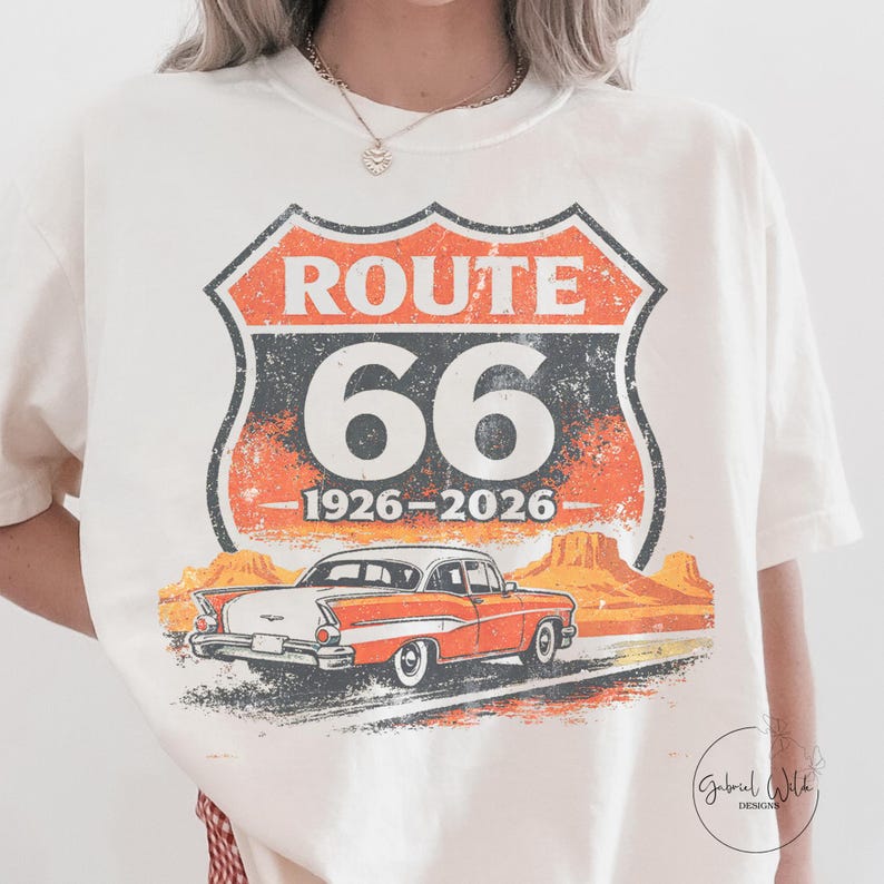 May include: Cream-colored t-shirt featuring a Route 66 graphic. The design includes a vintage car, the iconic Route 66 shield, and the text "ROUTE 66 1926-2026". The graphic is in shades of orange, black, and white.