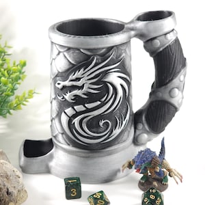 May include: A silver dragon-themed dice tower with a black handle and a separate dice tray. The tower has a dragon design on the front and a scaly texture. There are four green dice with gold numbers on a white surface.