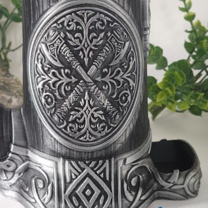 May include: A silver metal dice tower with a detailed design of crossed axes and floral patterns. The tower has a blue plastic dice tray and a decorative base.