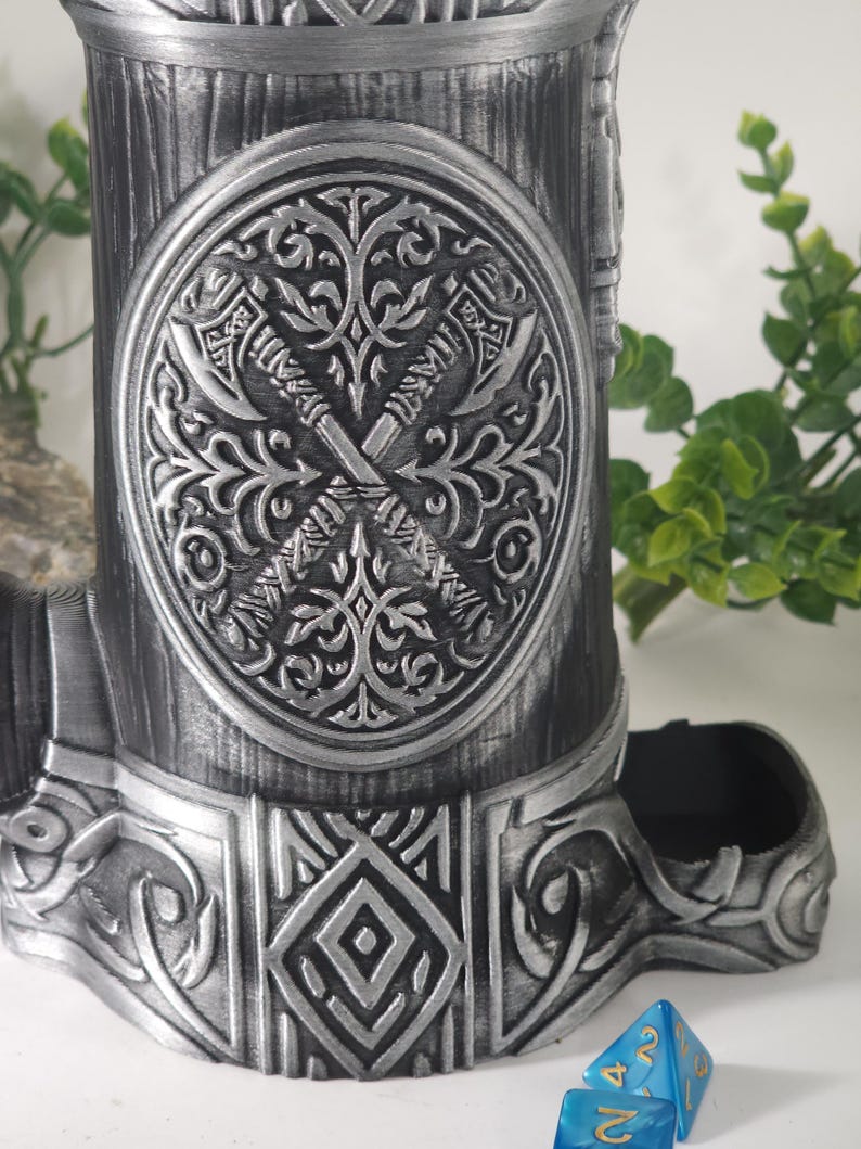 May include: A silver metal dice tower with a detailed design of crossed axes and floral patterns. The tower has a blue plastic dice tray and a decorative base.