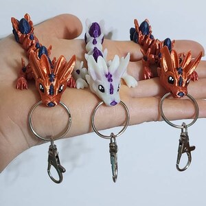May include: Three small dragon keychains with purple and white accents. The dragons are orange, white, and orange with black accents. Each keychain has a silver ring and a silver clip.