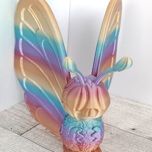 May include: A 3D printed butterfly figurine with iridescent wings in shades of blue, purple, and gold. The butterfly has a detailed, textured body and large, round eyes.