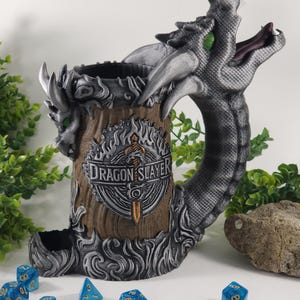 May include: A silver dragon head mug with a wooden barrel body. The mug has a dragon slayer logo with a sword and flames. The mug is surrounded by blue dice with gold numbers.