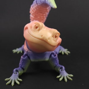 May include: A small, colorful, articulated gecko figurine with a pink body, purple legs, and a green and purple tail. The gecko has a friendly expression with large eyes and a slight smile.
