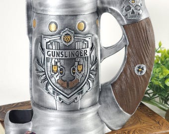 May include: A silver metal beer stein with a gun-shaped handle and a skull on the top. The stein has a crest with the word "GUNSLINGER" on it. The stein is sitting on a brown leather-bound book with dice scattered around it.