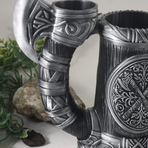 May include: A silver metal mug with an axe handle. The mug has a wood-like texture and a circular design with a stylized axe and floral patterns. The axe handle is wrapped in a silver band with a knot design.