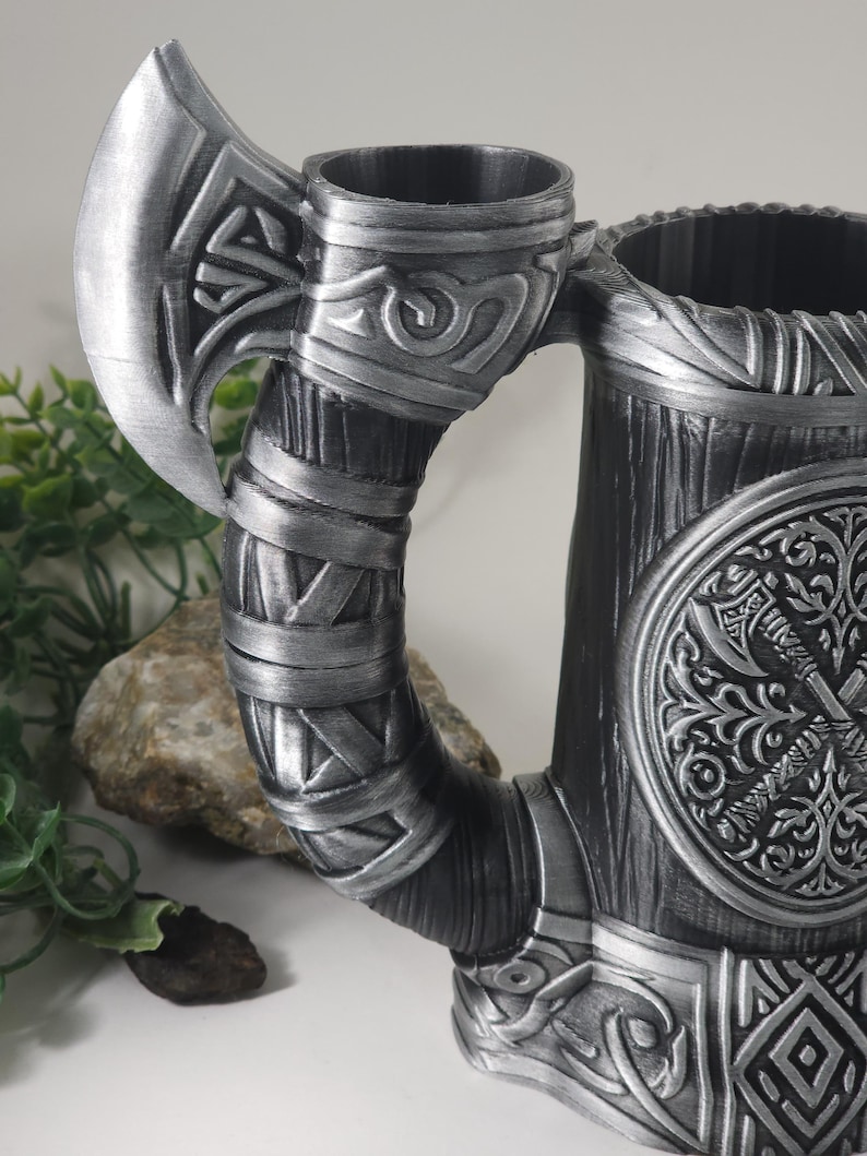 May include: A silver metal mug with an axe handle. The mug has a wood-like texture and a circular design with a stylized axe and floral patterns. The axe handle is wrapped in a silver band with a knot design.