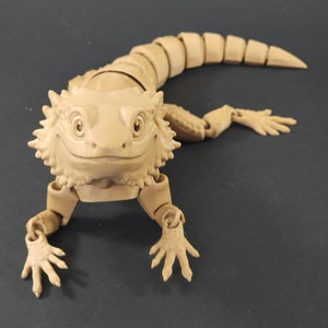 May include: A beige articulated figurine of a bearded dragon lizard with a friendly expression. The figurine is posed in a crawling position with its head raised and its limbs extended.
